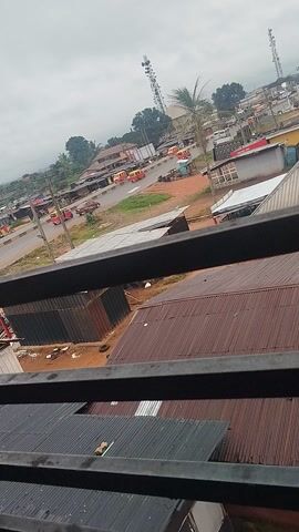 This video shows a street scene in Uselu, Nigeria, viewed from a high vantage point, possibly a balcony, as indicated by the presence of metal bars in the foreground. The scene is dominated by a road with several vehicles, including numerous red and yellow auto-rickshaws (Keke-NAPEPs), suggesting active transportation. Several buildings line the street, featuring corrugated metal roofs, common in many tropical regions. A large communication tower is visible in the distance, a common feature in urban and semi-urban areas of Nigeria. The sky is overcast, indicating a cloudy or possibly rainy day, typical of the weather patterns in Nigeria. The general atmosphere appears to be that of a busy market day or a typical weekday with commercial activity. The presence of multiple Keke-NAPEPs suggests a bustling local economy and transportation system. There is no discernible text in the video. The time of day appears to be daytime, given the level of natural light, but the cloudy conditions make it difficult to pinpoint the exact hour. The overall impression is a lively street in a Nigerian town.