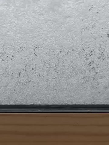 The video shows a window being opened to reveal a snowy scene outside. Initially, the window is opaque with snow. The window's latch is turned, and the window swings open, showing rooftops covered in snow, a yard, and a street. It is snowing. The window is then closed again.