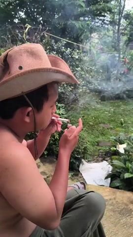 A bare-chested man in a tan cowboy hat is squatting on an outdoor surface in a garden setting, smoking a cigarette. He lights the cigarette, takes a drag, and exhales smoke into the air.  The garden behind him is lush with green plants and some flowers, with a glimpse of a house in the background.  He is wearing green pants and grey sandals.  He holds the cigarette in his hand while exhaling, then brings the cigarette back to his mouth for another drag.