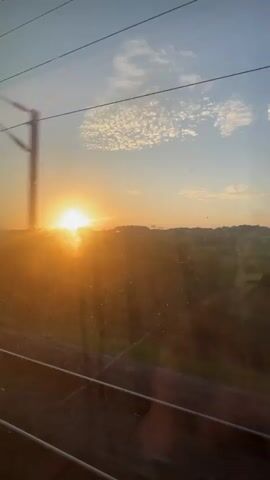 The video shows a view from a moving vehicle, likely a train, looking out a window. The sun is low in the sky, appearing to set or rise, casting a warm, golden light across the landscape. The sky is blue with scattered white clouds. Below the sun, there is a distant horizon line with rolling hills and some greenery. In the foreground, there are overhead wires and railway tracks, indicating the vehicle is on a train. The window itself has some smudges and reflections, one of which briefly shows the side of a person's face. A large tree is seen in motion, blurring as the vehicle passes by. The overall impression is of a scenic journey during the golden hour.