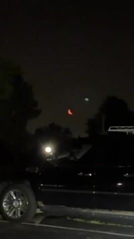 The video shows a night sky with a crescent moon, which appears orange. There are silhouettes of trees and possibly buildings in the foreground. A bright light, possibly a streetlamp, is visible in the lower left corner of the frame. The sky is dark and appears slightly hazy.