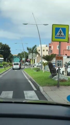 The video captures a view from inside a vehicle driving down a paved road. A large green vacuum truck is visible in the distance, traveling in the same direction. To the right of the road, there is a well-maintained grassy area with a palm tree and several street signs. One sign indicates directions to "Túnez," "La Camella," "La Sabinita," and "Arona." Other signs include a yield sign, a pedestrian crossing sign, and a warning sign with children depicted. A white truck is parked on the side of the road. Buildings with apartments and shops line the right side of the street. The sky is partly cloudy.