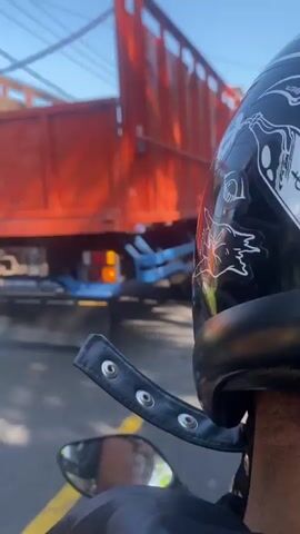 The video shows a first-person perspective of someone riding a motorcycle. The rider is wearing a black helmet with white decals of skulls and other illustrations. Part of the rider's neck and shoulder are visible, along with a black jacket. In the background, a large orange truck with a blue chassis is passing by. The road has a yellow line marking the lane. Further ahead, trees, houses, utility poles, and wires can be seen under a clear blue sky. The motorcycle has a visible rearview mirror. The overall environment suggests a sunny day on a road, possibly in a rural or suburban area.