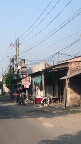 A street scene on a clear day. A young man on a motorcycle is seen from the side as he rides along a paved road. He is wearing a dark shirt and dark pants. The road is lined with small shops. One shop appears to be a repair shop with tools and equipment visible. Another shop has a sign that says "TAMBAL BAN" which means tire repair. There is a bicycle leaning against a building. Power lines are visible overhead. The overall impression is of a rural or semi-rural environment.