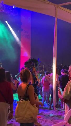 The video shows a crowd of people at an outdoor event with a sign that says "End of SUMMER". A person in a large, colorful feather headdress and blue bikini is dancing and interacting with people in the crowd. There are stage lights illuminating the scene with blue, green, and pink lights. Many people in the crowd are dressed casually, and some are recording with their phones. The event appears to be taking place on a beach under a tent or canopy.