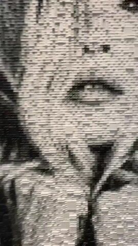 The video captures a slow zoom-out of a large-scale, monochromatic mosaic artwork depicting the close-up face of an infant or toddler. The artwork is composed of countless small, rectangular tiles in black, white, and various shades of gray, creating a textured, pixelated image. The child's face, rendered in grayscale, looks directly forward. The scene is indoors, likely an exhibition or gallery setting. The video was filmed in Noordwijk, Netherlands.
