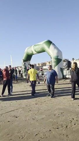 The video shows a group of people gathered on a beach. The crowd includes people in wetsuits, some wearing swimming caps, seemingly preparing for a swimming event. There's a large inflatable arch marking the start or finish line of the event. The beach itself appears sandy, and the sky is clear and blue. Some people are standing around talking or stretching, while others are walking towards the water. The atmosphere seems relaxed and anticipatory. In the background, some buildings and possibly a port area can be seen.