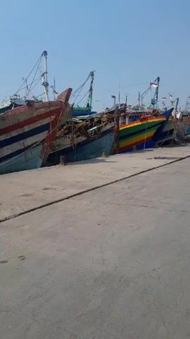 The video shows a dock with several large fishing boats moored alongside. The boats are of various sizes and colors, with some displaying multiple stripes or patterns. The sky is clear and blue, indicating a sunny day. The ground is a paved surface, likely concrete, with a thick rope running across it. The boats appear to be either docked or under repair, with some showing signs of wear and tear. One boat in the middle has a rainbow-colored hull. The video pans across the scene, providing a view of the boats from different angles. There are no visible people or distinct actions occurring in the video, other than the slight movement of the camera.