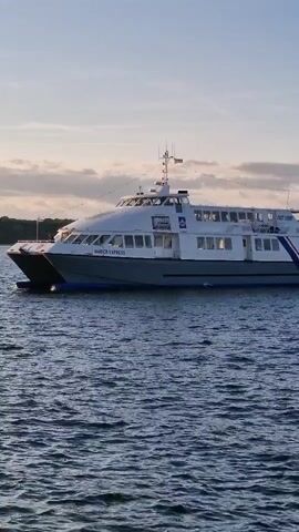 A passenger ferry with the name "MAVERICK EXPRESS" printed on its side moves across choppy, dark blue water. The ferry has a white upper deck with numerous windows and a grey hull. The sky above is a pale blue with scattered clouds, and a tree-lined shore is visible in the distance. The sun appears to be low on the horizon, casting a soft light on the scene.