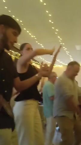 This video shows a group of people dancing indoors. There are string lights draped across the ceiling. In the foreground, a man with a beard and a woman in a black top and white pants are dancing. The woman is holding a large, round, white object. Behind them, a man in a light blue shirt and khaki pants is dancing, and another man in a white shirt and light green pants is also dancing. Further back, a woman in a red dress is dancing with another person, and a woman in a dark dress is visible. The floor appears to be wood. The lighting is warm and yellow.