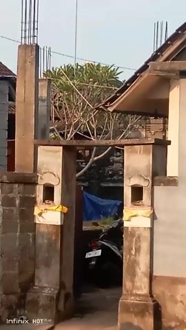 The video shows a close-up view of a stone gateway with two pillars. Each pillar has a small recessed niche at the top, decorated with a yellow cloth and what appear to be offerings. Beyond the gateway, a motorcycle is parked and a blue tarp is visible. In the background, there are trees and part of a building with a pitched roof. The sky is visible, suggesting an outdoor setting. The video is static, with no visible movement within the frame.