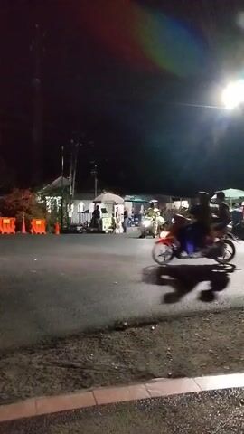 A nighttime scene shows a street with several motorcycles and people. The road is paved, and there are structures along the side, possibly shops or stalls. A street light illuminates the area. One motorcycle in the foreground has two individuals riding it. Other motorcycles and people are visible in the background, some of whom appear to be interacting. The lighting suggests it is dark, with the street light being the primary source of illumination. Orange barriers are present on the left side of the road. The overall atmosphere appears to be lively, possibly a market or gathering.