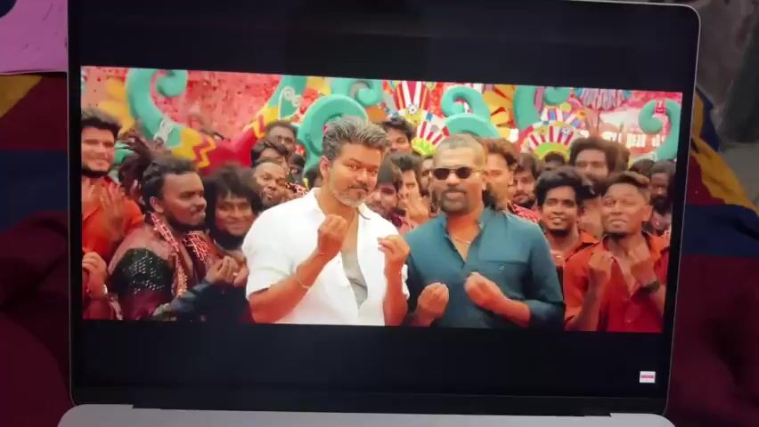 A laptop screen displays a music video featuring a group of men dancing in an outdoor setting. Two men in the foreground, one in a white shirt and the other in a dark blue shirt and sunglasses, are gesturing with their hands and smiling. They appear to be the central figures of the scene. Behind them, a large group of men, dressed in colorful traditional Indian attire, are energetically dancing and moving in unison. The background is brightly decorated with vibrant banners and colorful structures, suggesting a festive atmosphere. Red powder or paint is being thrown or sprayed, creating a dynamic and colorful visual effect. The scene captures a lively and celebratory moment from the video.