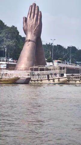 The video shows a large bronze statue of two hands clasped together in a prayer or greeting gesture, positioned in a body of water. The statue is situated on what appears to be a raised platform or embankment. Several boats are docked or moving in the water in front of the statue, with people visible on the boats. The background shows a shoreline with trees and buildings, and a cloudy sky. The camera is positioned at water level, looking towards the statue and the boats.