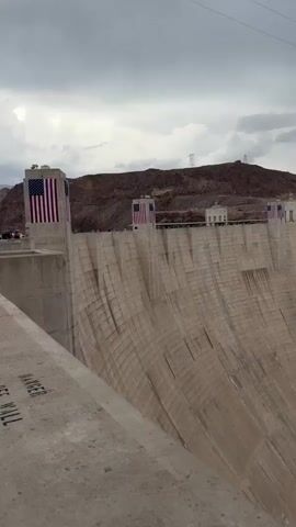 A wide-angle, vertical video pans across the face of a massive concrete dam under a cloudy sky. Several American flags are visible on top of the dam, as are small white buildings and structures. The dam's surface is textured and appears to be composed of many individual concrete sections. To the right of the dam, rugged, reddish-brown rock formations rise steeply, with power lines and pylons visible against the sky in the distance. The camera movement is slow and deliberate, giving a sense of the immense scale of the dam and the surrounding landscape.
