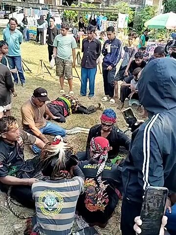 The video shows a group of people gathered around a figure lying on the ground in an outdoor setting. Several individuals appear to be attending to the person on the ground, with some kneeling or sitting close by. A few are dressed in what appears to be traditional or performance attire, indicated by face paint and specific clothing. Others are standing around, observing the scene. The environment suggests a grassy area with buildings visible in the background. Many people are holding or looking at their phones, possibly recording or documenting the event. Overall, the scene captures a moment of interaction and attention focused on the individual on the ground, surrounded by onlookers and helpers.
