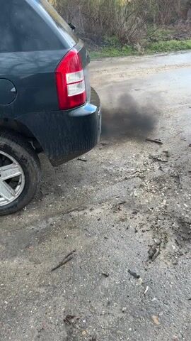 A dark gray hatchback car is parked on a muddy, uneven road next to a grassy, wooded area. The car is dirty, with mud splattered on the lower portion of the vehicle. The rear tire is partially visible. The road is covered in mud, twigs, and small debris. The car's engine is running and emitting a puff of white smoke from the exhaust pipe. A yellow building is visible in the background. The overall lighting is overcast, suggesting a cloudy day.