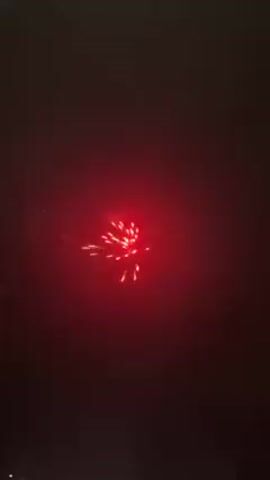 The video depicts fireworks exploding in the night sky.  Multiple fireworks are launched, each creating distinct bursts of light with varying colors including red, orange, and yellow. The sky is dark with some cloud cover visible.