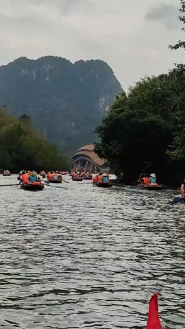 The video shows a river scene with numerous orange boats, each carrying several people wearing orange life jackets, moving through the water. Some people are actively rowing with oars. In the background, a large mountain looms behind a building with a thatched-style roof. Trees are visible on either side of the river. The sky is overcast. The scene appears to be a tour on a popular river route.