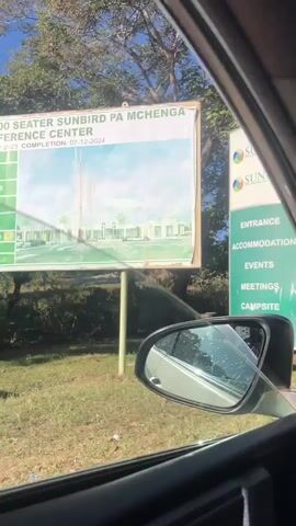 The video, shot from inside a car, shows a roadside sign for Sunbird Waterfront and Sunbird Livingstonia, listing entrance, accommodation, events, meetings, campsite, and watersports. The car moves forward, revealing a guard booth or entrance gate ahead. Stickers are visible on the car's windshield. The sky is blue with sunlight. The car's interior is visible, including the side mirror, dashboard, and speaker. Trees and foliage are visible in the background.