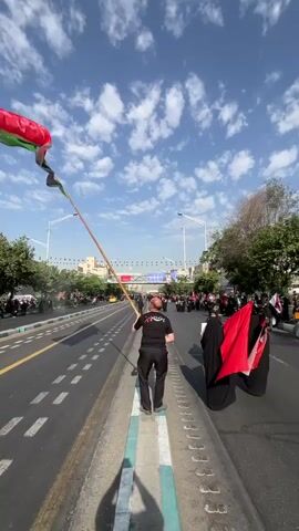 A man in a black t-shirt and black pants holds a long wooden pole with an Iranian flag at the top. He is standing on the side of a wide road, facing away from the camera, with a crowd of people gathered behind him. The flag is waving in the wind. There are cars and buildings in the background, and the sky is blue with some clouds. The people behind him are dressed in dark clothing, and some are carrying red flags. The scene appears to be a procession or demonstration.