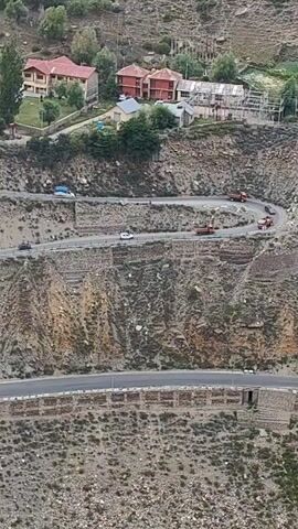 The video displays a winding mountain road with several vehicles traversing it. The road is carved into a steep, rocky mountainside. Several buildings are visible at the top of the mountain, and the terrain is characterized by rugged slopes and sparse vegetation. The vehicles include cars, trucks, and what appears to be a delivery van or small truck. The road has guardrails in some sections, and retaining walls made of stone are present to prevent erosion. The overall scene depicts a challenging but functional road system in a mountainous region.