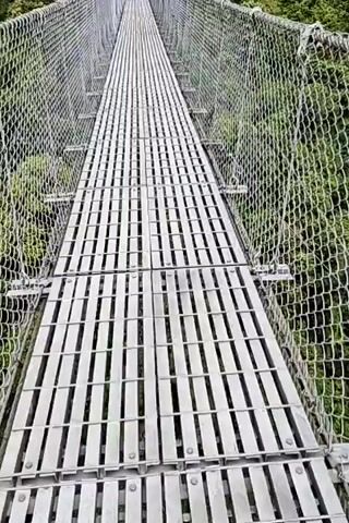 The video shows a long, narrow suspension bridge made of metal grating and wire mesh. The bridge spans across a lush, green forested valley, with steep, tree-covered hillsides on either side. The camera appears to be positioned at the beginning of the bridge, looking down its length towards a distant point where it disappears into the trees. The bridge seems to be suspended high above the ground, though the depth is not clearly discernible. The overall impression is one of great length and height, emphasizing the natural beauty of the surrounding landscape.