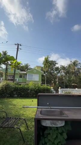 The video shows a view of a sunny outdoor area with a green lawn, palm trees, and several buildings, including a green house and a partially constructed building behind a white picket fence. A swimming pool is also visible. The camera then moves to show a body of water, possibly a lake or river. Finally, a young woman appears in the frame, sitting by the water. She is wearing headphones, a t-shirt, and shorts, and she makes a peace sign at the camera.