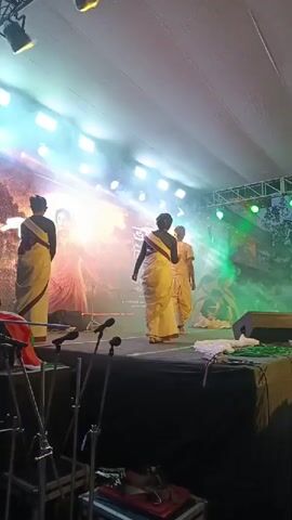 The video shows a stage performance. There are four people on the stage, dressed in traditional Indian attire. They appear to be performing a dance or a play. The stage is lit with bright lights, and there is smoke in the air, creating a dramatic effect. In the background, there is a large screen displaying an image. In the foreground, there are microphones and stage equipment. The overall atmosphere is festive and engaging.