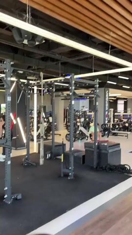 A wide-angle shot shows the interior of a modern gym. In the foreground, a black squat rack with pull-up bars stands on a black rubber floor. Other equipment like TRX straps and plyometric boxes are visible around the rack. In the background, other gym-goers can be seen using various exercise machines, including treadmills and weight machines. The gym features a contemporary design with exposed ceilings, wooden accents, and bright linear lighting. A sign visible on a back wall reads "CROCODILUS FITNESS". The overall impression is a well-equipped and spacious fitness facility.
