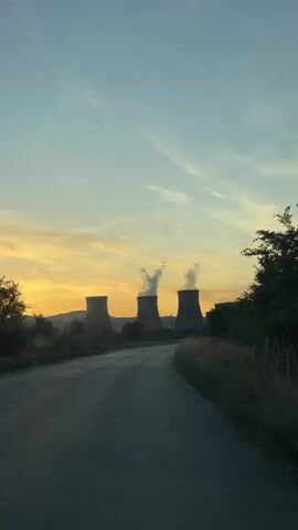 The video shows a view from a vehicle moving along a road toward a power plant with cooling towers. The sky is clear with some wispy clouds, and the sun is setting, casting a warm orange glow on the horizon. The cooling towers are emitting steam, creating plumes that rise into the air. The road is paved and appears to be empty. The surrounding landscape is mostly rural, with some trees and dry grass visible on the sides of the road. The overall impression is one of industrial activity set against a natural landscape at dusk.