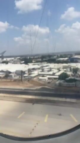 The video shows a view from an airplane window looking out at a runway and the surrounding landscape. The runway is made of concrete with yellow lines marking its edges and a portion of the center. The area beyond the runway contains several large, white-roofed buildings, some trees, and a road with what appear to be vehicles. In the distance, there is a hazy view of more buildings and landscape under a partly cloudy blue sky. The airplane wing is visible on the left side of the frame. The camera appears to be stationary.