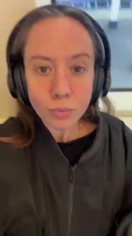 A woman wearing headphones and a black jacket looks at the camera and rubs her right eye with her right index finger. She then makes a kissing face toward the camera.