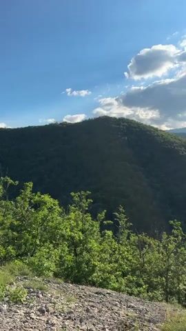 The video pans across a lush green mountainous landscape under a partly cloudy blue sky. The sun is visible, shining brightly and casting a warm glow. In the foreground, there are trees and rocky ground. The mountains are covered in dense vegetation, with rolling hills and valleys creating a sense of depth and scale. The overall impression is one of natural beauty and tranquility.