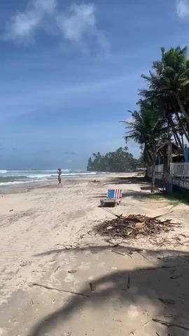 A vertical, full-shot video captures a beach scene under a bright, cloudy sky. The foreground is dominated by a sandy beach with scattered debris. A single, rustic chair adorned with an American flag design sits near the debris. In the midground, a person stands by the ocean's edge, facing the water, appearing to hold something in their hands. The ocean features moderate waves crashing onto the shore. The background reveals a lush coastline with numerous palm trees and a dense jungle. A small, rustic structure with a white railing and a sign that reads "LIFE GUARD" is visible to the right of the frame. The overall ambiance is one of a peaceful, tropical day at the beach.
