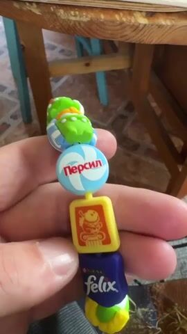A hand holds a string of connected plastic beads. The beads are mostly green and represent various brand names and cartoon characters. The bottom bead depicts a Felix brand cat food product in blue, yellow, and green. Above that, a yellow rectangular bead features an image of a character. Above that is a blue and white round bead with the word "Persil" printed on it. The remaining beads depict green cartoon characters with various colored hats. The background is blurry and shows a wooden table and a patterned rug.
