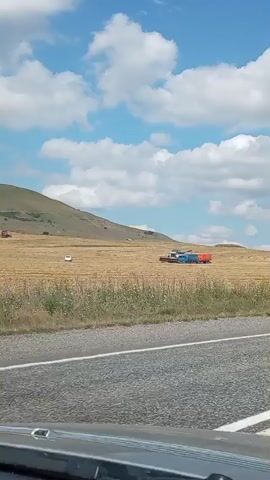 A wide shot shows a rural landscape with a field of dry grass under a partly cloudy sky. In the distance, a red tractor is visible to the left, and a blue combine harvester is working in the field to the right, pulling an orange trailer. A white car is parked in the field near the tractor. The foreground shows the edge of a road, suggesting the footage is being recorded from a vehicle.
