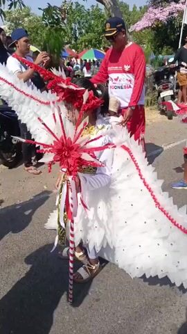 The video captures a festive outdoor event with a parade. In the foreground, a child is dressed in an elaborate costume resembling a bird with large white feathered wings and a red and white headdress. The child holds a decorative staff adorned with red and white ribbons and a large red floral ornament. To the left and behind the child, a young person in a white shirt and blue cap is adjusting greenery. To the right and behind the child, a man in a red shirt with a white design and a dark cap is standing, looking at the child. Further in the background, there are other people, including children, and parked motorcycles. A red and white Indonesian flag is visible on a pole in the upper right portion of the frame, and trees with pink blossoms are present in the background. The ground appears to be a paved road. The overall atmosphere suggests a celebration or cultural event.