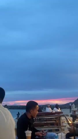 The video shows a group of people sitting at tables on a sandy beach. The sky is a deep blue with some pink and orange hues from a sunset. In the foreground, a man in a black shirt is looking down at his phone, with a coffee drink on the table in front of him. To his right, another person is holding up a phone, possibly taking a picture or video. Further back, several other people are seated at tables and walking along the beach. There is a body of water with a rocky breakwater in the distance. The overall atmosphere appears relaxed and casual.