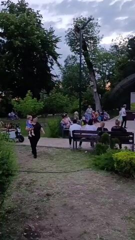The video shows an outdoor gathering in a park-like setting. In the foreground, a dirt path runs diagonally from the bottom left to the bottom right, with a paved walkway bordering its right side. To the left of the path is a grassy area with some bushes. In the midground, people are seated on benches facing a stage area. Several people, primarily adults and some children, are gathered on and around the stage, with some appearing to be dancing. A banner with text and a rainbow graphic is visible on the stage. Surrounding the stage and seating area are numerous trees, indicating a wooded environment. The sky above appears overcast, suggesting it is either early morning or late evening. The overall atmosphere seems casual and social, with a performance or activity taking place on the stage.