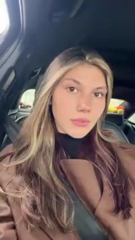 A young woman with long blonde and brown hair is in the driver's seat of a car. She wears a brown coat over a black turtleneck. Her seatbelt is fastened. She looks directly at the camera and nods slowly. The interior of the car appears dark. A small yellow stuffed animal is in the back seat. Music plays audibly.