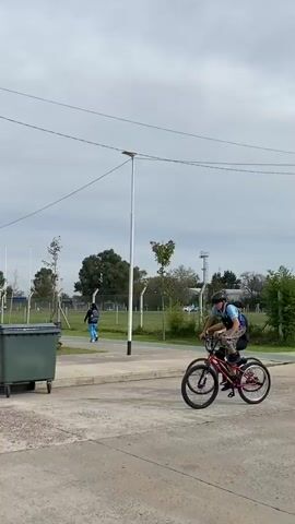 The video shows a young man wearing a helmet, a blue t-shirt with camouflage shorts and black shoes riding a red BMX bike on a paved road in a park.  He is standing on the frame of the bike, with his feet on the seat and handlebars, propelling it forward. Another young man is riding nearby, also on a bike. In the background, there is a green lawn with palm trees, park lights, and a couple of people walking. A tall metal structure, possibly a cell tower, stands in the background.  There is also a large green waste container in the foreground. The sky is overcast.