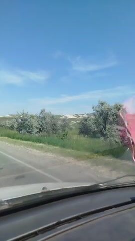 The video shows a view from inside a car driving down a paved road on a sunny day. The sky is clear and blue with a few wispy clouds. The road is bordered by green grass and trees. In the distance, there are rolling hills. There are no other vehicles or people visible in the video. The driver's dashboard is partially visible in the foreground.