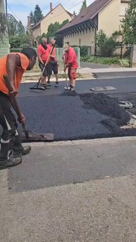 Four construction workers are resurfacing a road. Two workers are in the foreground, spreading asphalt with shovels. One worker is wearing an orange shirt and black pants, and the other is wearing a red shirt and red pants. In the background, two other workers in red shirts and red pants are standing near a paving machine. The road is partially covered with fresh asphalt, and there is a pile of asphalt on the side of the road. The scene appears to be in a residential area, with houses and trees visible in the background.