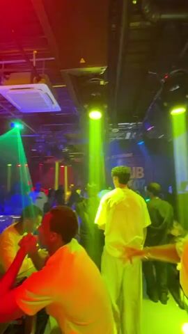 The video shows the inside of a club with many people and colorful stage lighting.  The camera pans slightly from left to right, offering different viewpoints of the crowd. Some people are standing around in groups, and a couple of individuals sit near a bar, while a DJ appears to be performing on a raised platform in the background. The club is brightly lit with colored stage lights, which creates an atmosphere of activity and music.