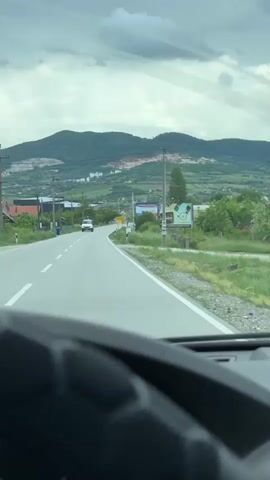 The video, shot from inside a vehicle, depicts a view of a straight road leading into a town in a rural setting. The camera focuses on the asphalt road with white lines marking the lanes. A white van with an IVECO logo is driving towards the camera. On the right side of the road, a yellow town sign labeled "ВРБИЦА" with Cyrillic script is visible. Numerous billboards also stand along the roadside. The surrounding environment includes green fields, trees, houses, and power lines running along the road. In the background, a large mountain rises, showing signs of quarrying or mining activity with exposed rock. The sky is overcast with grey clouds.