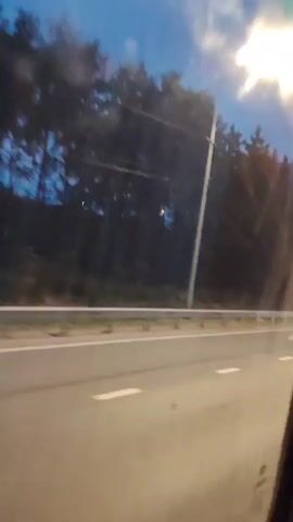 The video, filmed at dusk, shows a car driving on a highway at night. Trees line the side of the road, and the sky is a deep blue. The car's headlights are on, illuminating the road ahead. The camera is positioned inside another vehicle, looking out the window. The video appears to be a dashcam recording, capturing the perspective of a passenger.