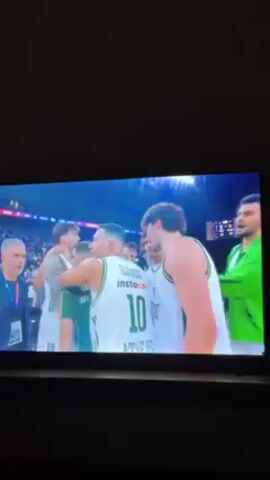 The video shows a basketball game on a television screen. In the first few seconds, players wearing white and green jerseys give each other high fives and clap hands. The person in the foreground is wearing the number 10 jersey. At the 3-second mark, the screen shows a graphic with the logos and names of the two teams, LDLC ASVEL and PANATHINAIKOS BC AKTOR ATHENS.