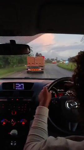 The video shows the interior view of a car as a person drives down a highway. A truck loaded with what appears to be bricks travels ahead. Other vehicles are visible in the distance. The sky is overcast with some hints of sunset colors. The car’s dashboard features illuminated gauges and controls, indicating the car is running. The driver's hand is visible on the steering wheel. Their curly hair suggests they are in the driver's seat.