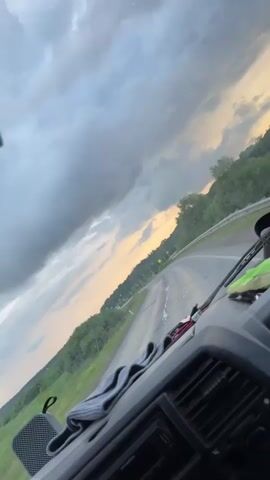 The video shows a view from inside a vehicle, looking out through the windshield. The vehicle appears to be driving on a paved road, which is wet. The sky is overcast with dark clouds, but there is a strip of light yellow and orange sky visible on the horizon, suggesting either sunrise or sunset. Trees and green foliage line the sides of the road. In the distance, there are a few cars on the road, one of which is a white SUV. There are also traffic signs visible along the road. The interior of the vehicle is partially visible, including a dashboard and a vent. The video has a slightly tilted perspective.